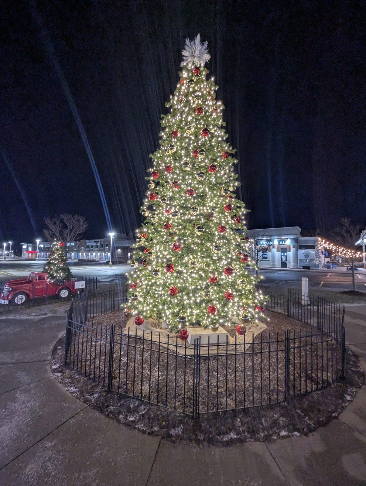 Two lit commercial Christmas trees and a vintage truck in a shopping plaza at night