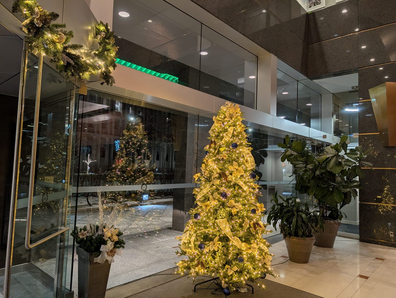 Illuminated Christmas tree and a lit wreath over the glass entrance of a modern corporate lobby.