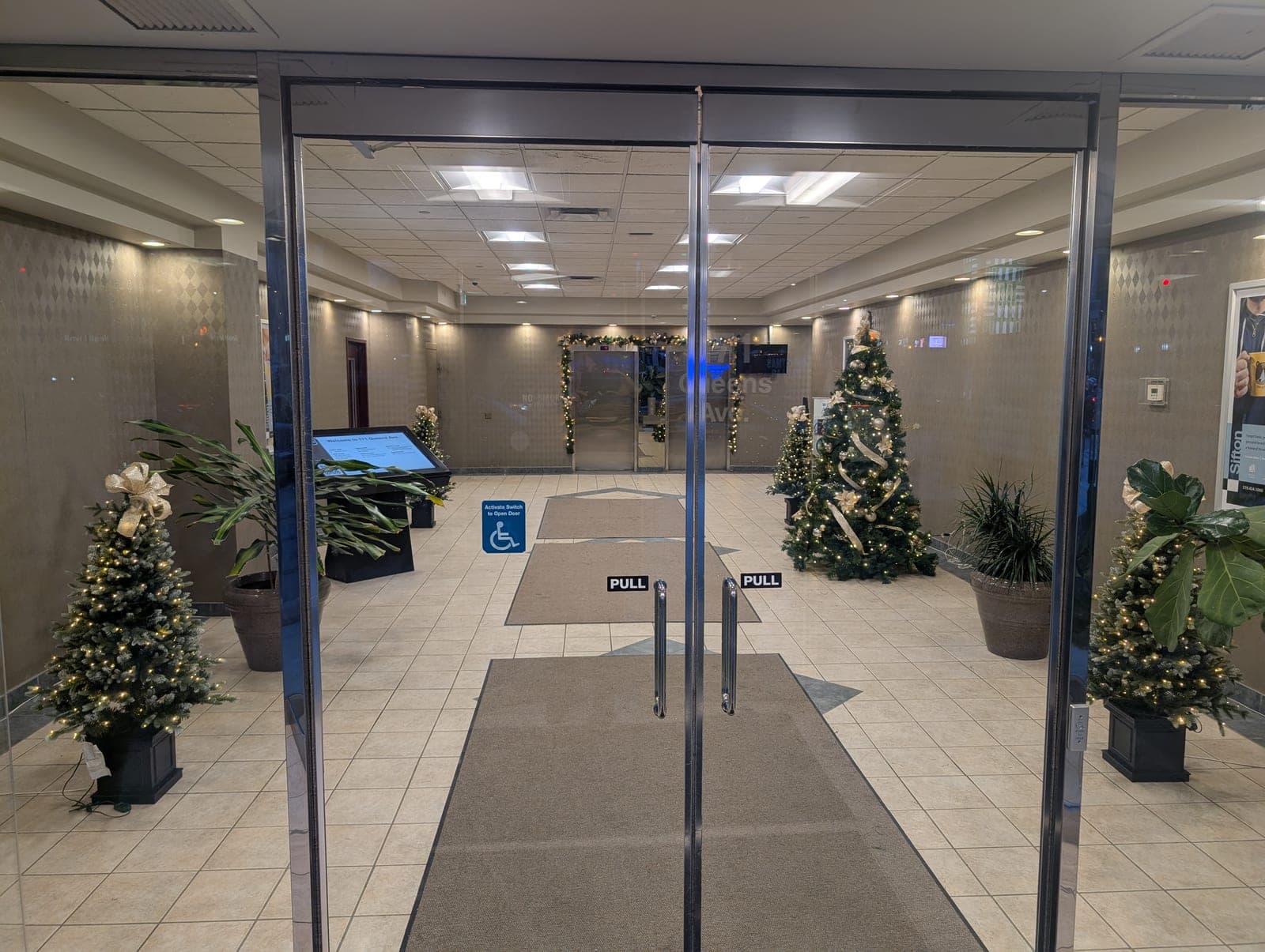 Commercial building lobby entrance flanked by lit Christmas trees and garland-wrapped interior doors