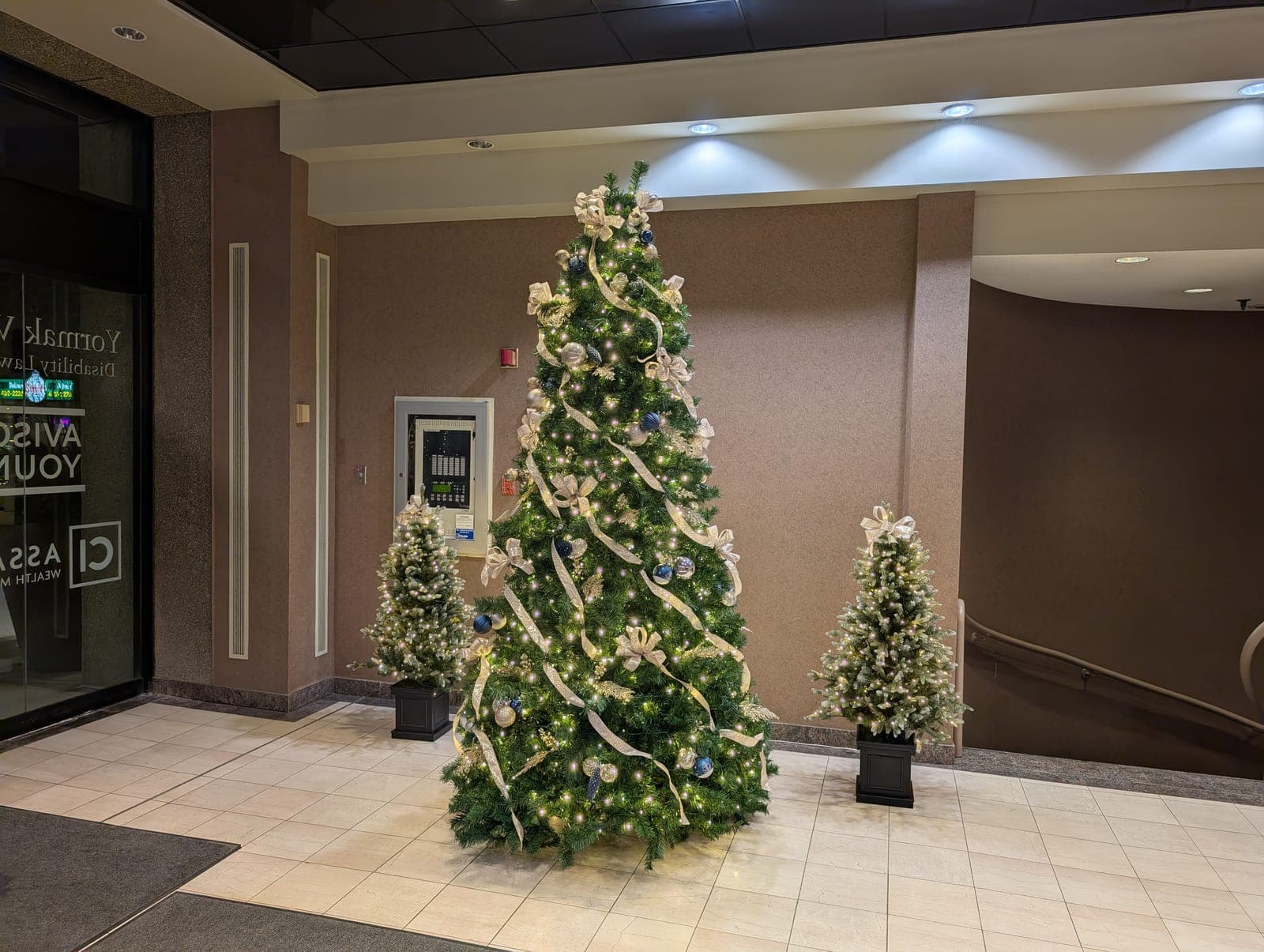 Corporate building lobby Christmas tree with gold ribbon flanked by two lit accent trees