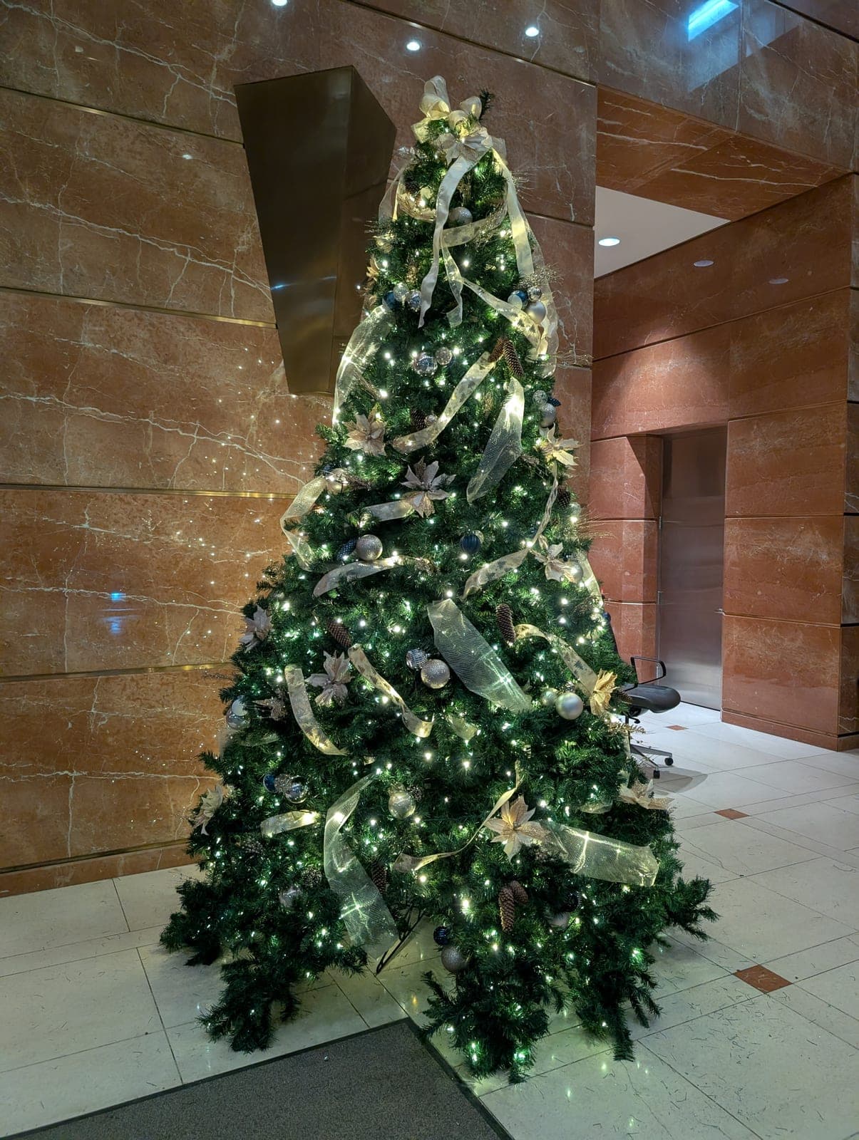 Decorated corporate lobby Christmas tree with gold ribbon and warm-white lights against a marble wall