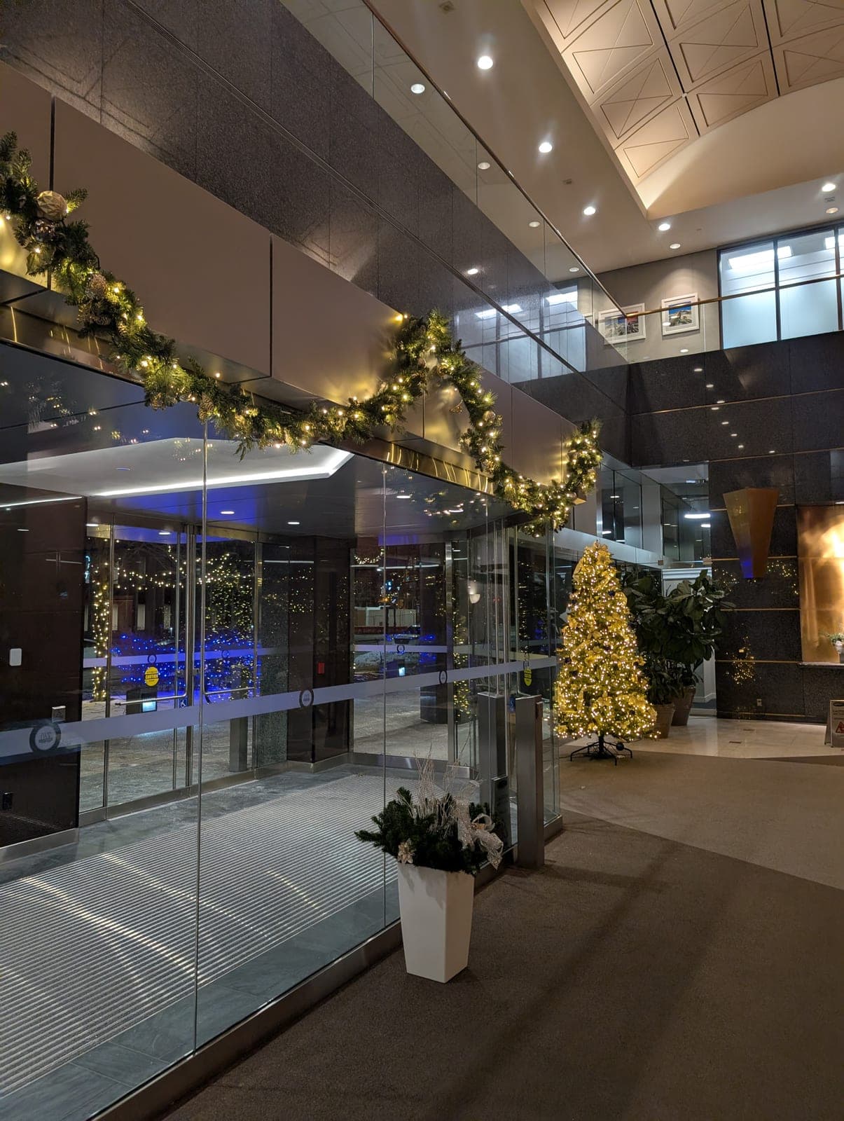 Lit evergreen garland draped over a glass vestibule and a warm-white Christmas tree in a corporate lobby.
