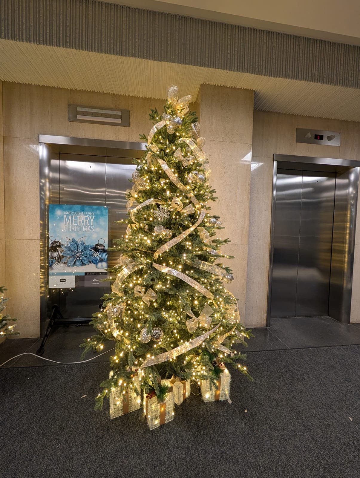 Decorated Christmas tree with gold ribbon and lit gift boxes between two office building elevators