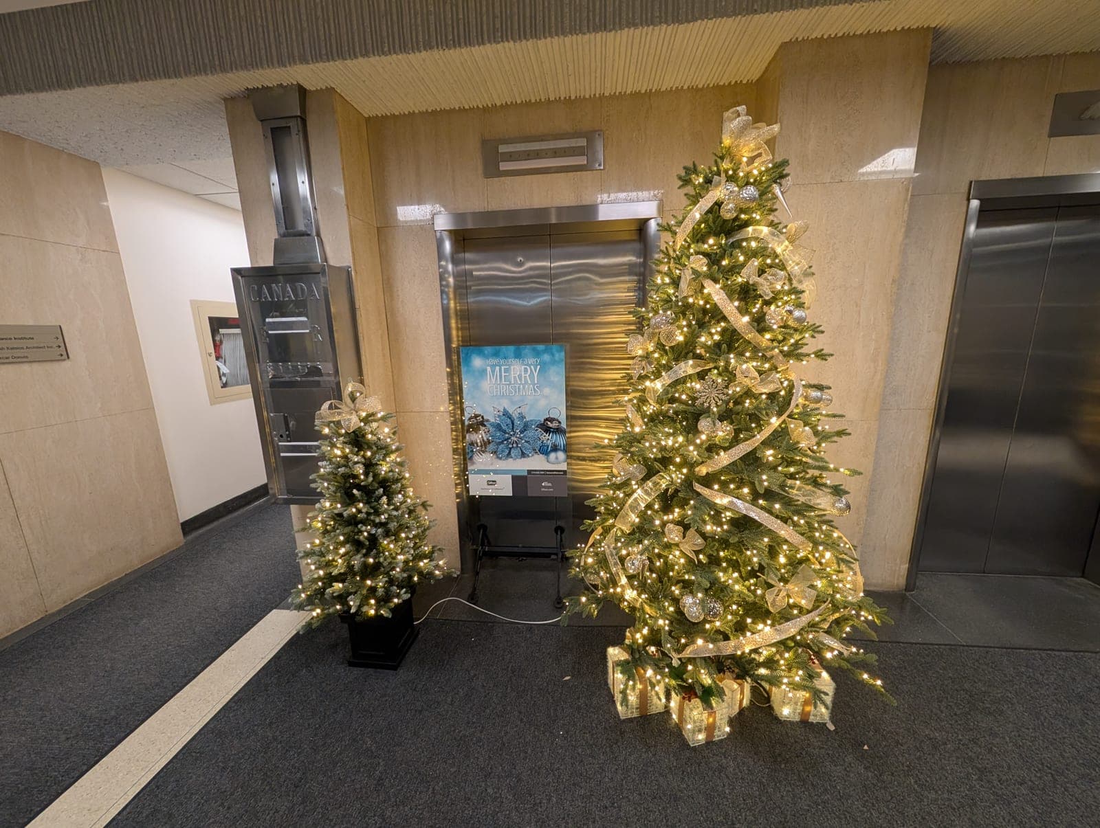 Gold-ribboned Christmas tree with lit gift boxes beside office elevators and a Merry Christmas sign