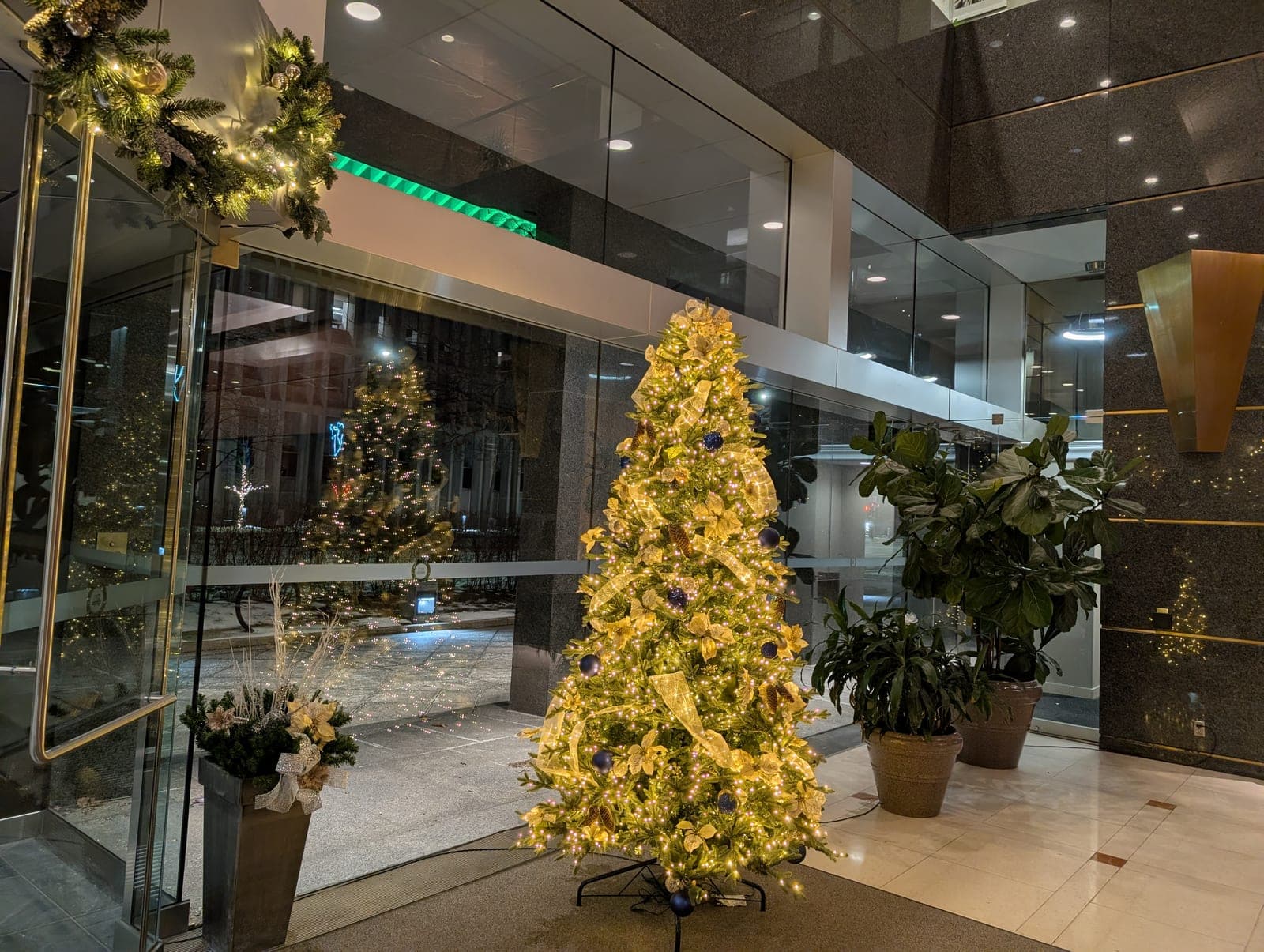 Warm-white lit Christmas tree with gold ribbon in a granite corporate lobby beside fiddle-leaf plants.